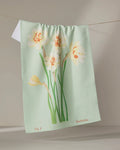 A light green recycled kitchen towel by Linda Fox, called "March Daffodils," features yellow and white daffodil illustrations. "Fig 3" and "Daffodils" are printed at the bottom as it hangs on a clothesline.