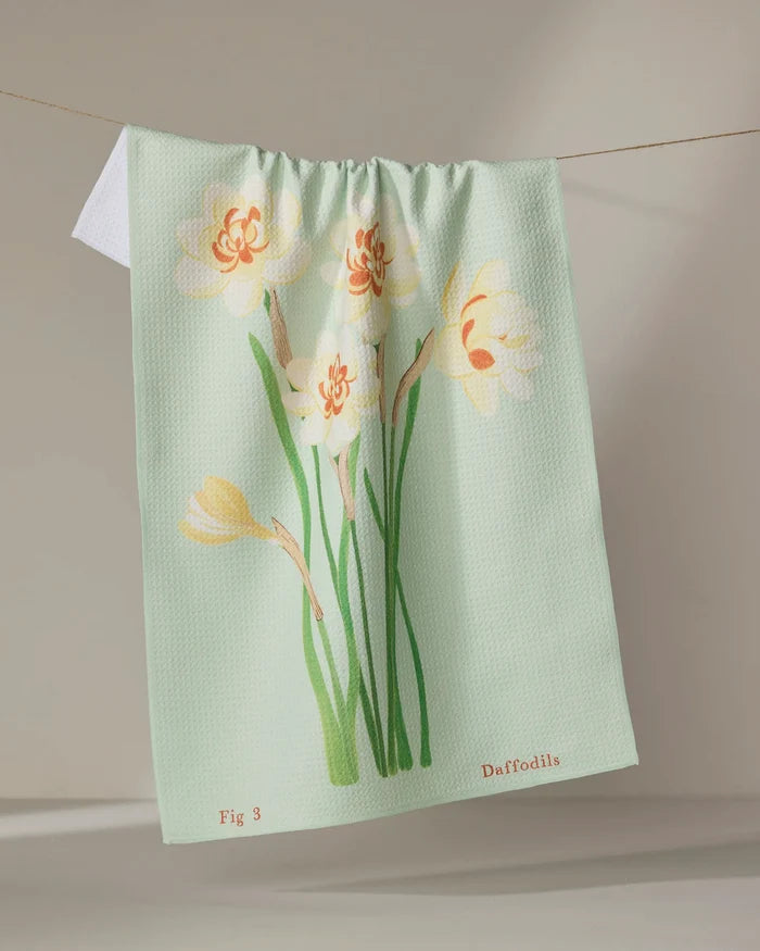 A light green recycled kitchen towel by Linda Fox, called "March Daffodils," features yellow and white daffodil illustrations. "Fig 3" and "Daffodils" are printed at the bottom as it hangs on a clothesline.