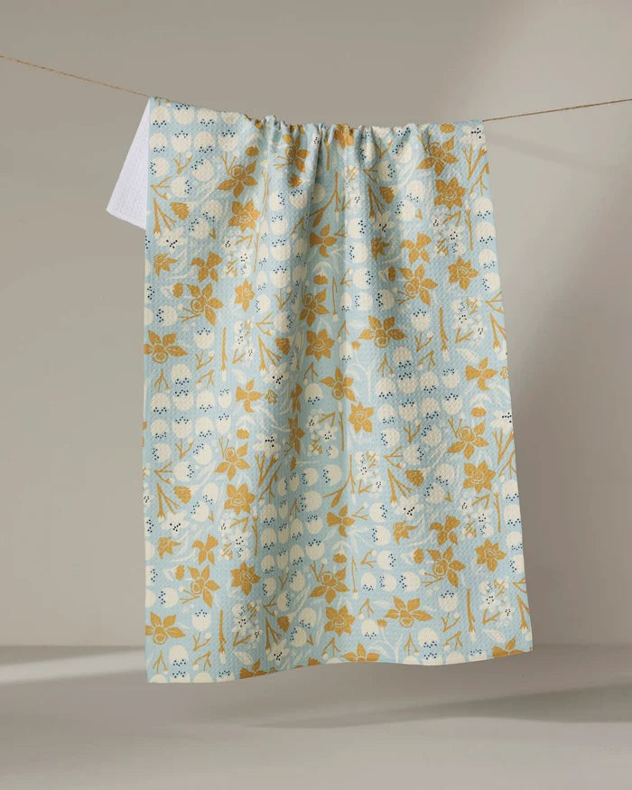 The Elisabeth Wing "March" tea towel, ultra-absorbent in light blue with a yellow floral and vine pattern, hangs on a clothesline against a neutral background.