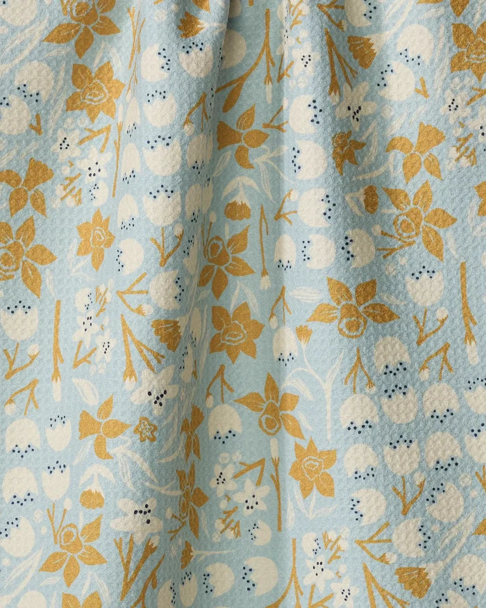 A close-up of the "March" tea towel by Elisabeth Wing, featuring light blue textured fabric adorned with white berries and golden yellow floral motifs for a soft, elegant look on this ultra-absorbent kitchen accessory.