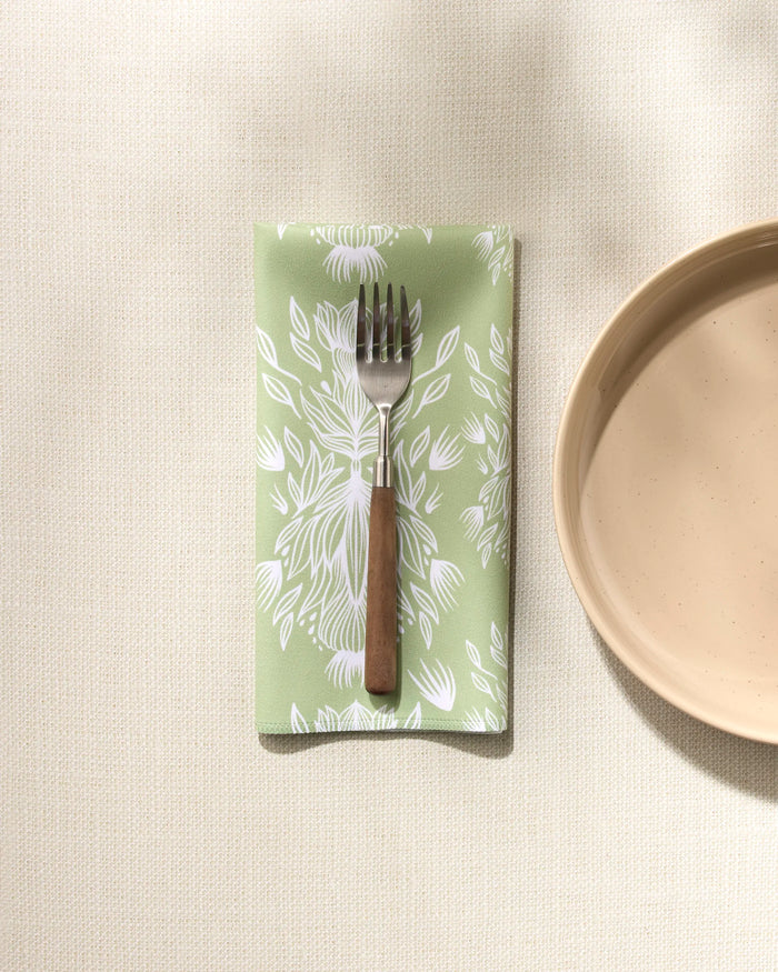 A GEOMETRY Margarita Vibes green reusable napkin with a white floral pattern sits folded on a light surface, holding a fork beside a beige ceramic plate. Sunlight casts gentle shadows across the scene.