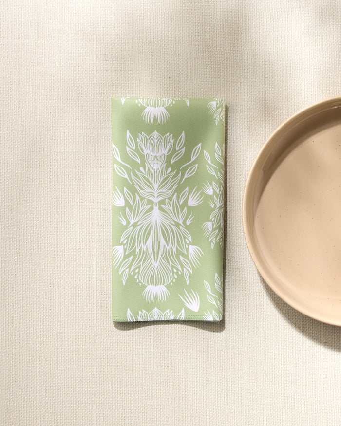 The GEOMETRY "Margarita Vibes" sage green reusable napkin with a white floral pattern rests on a textured cream tablecloth by a beige ceramic plate, bathed in soft natural light for an inviting look.