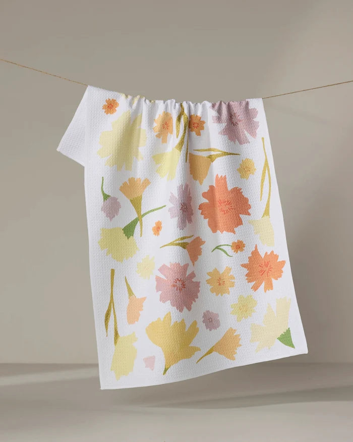 The Marigold Flowers tea towel by Elana Gabrielle features a textured white surface with pastel yellow, orange, green, and pink floral patterns. Made from recycled materials, it hangs on a clothesline against a neutral background.