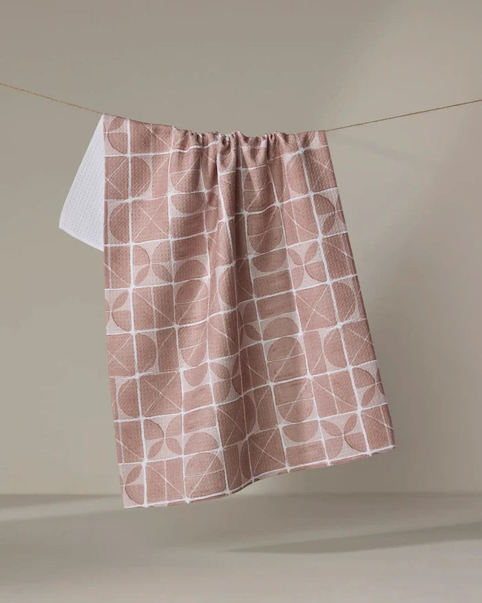 The Mariposa towel by Studio Jeannot, featuring a quick-drying beige fabric with a white geometric pattern, hangs from a string against a plain, neutral background.