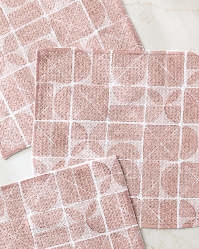Four Mariposa dishcloths by Studio Jeannot, in pink with a white geometric pattern, are arranged on a light marble surface.