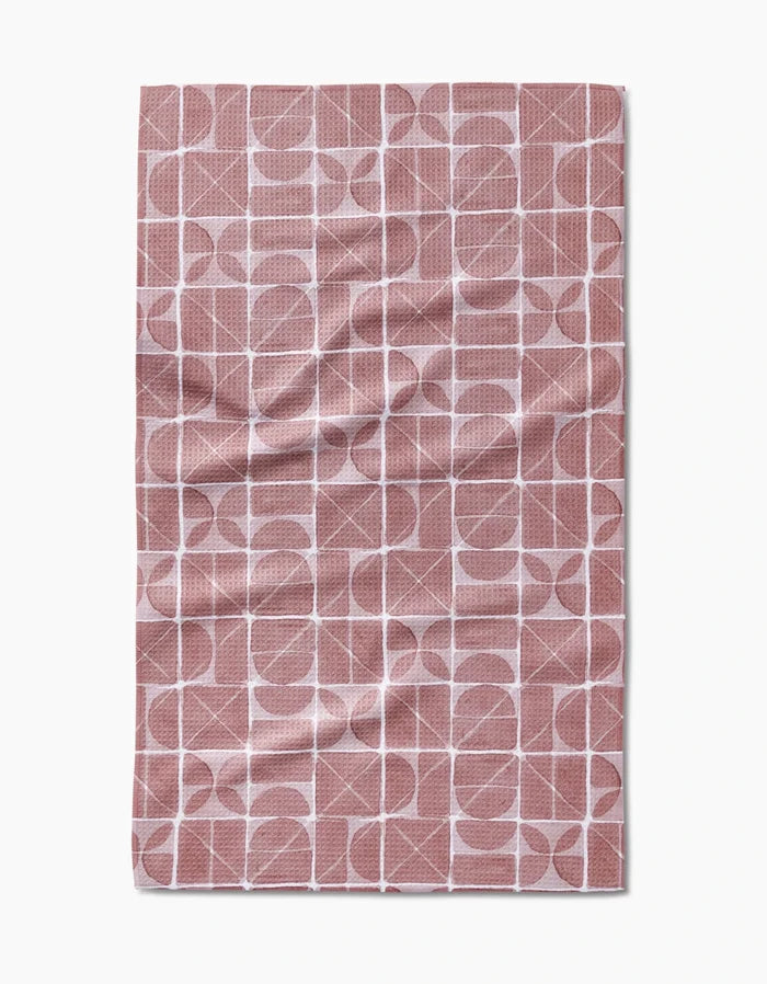 The Mariposa tea towel by Studio Jeannot is a rectangular kitchen towel with a pink and white geometric grid of circles, semi-circles, and squares. Ultra-absorbent, it features visible creases and is set against a white background.