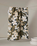 The Marlo Brown tea towel by Geometry, featuring a bold floral pattern in brown, beige, and white, hangs above a stack of white plates against a neutral background.