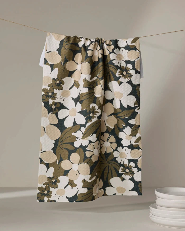 The Marlo Brown tea towel by Geometry, featuring a bold floral pattern in brown, beige, and white, hangs above a stack of white plates against a neutral background.