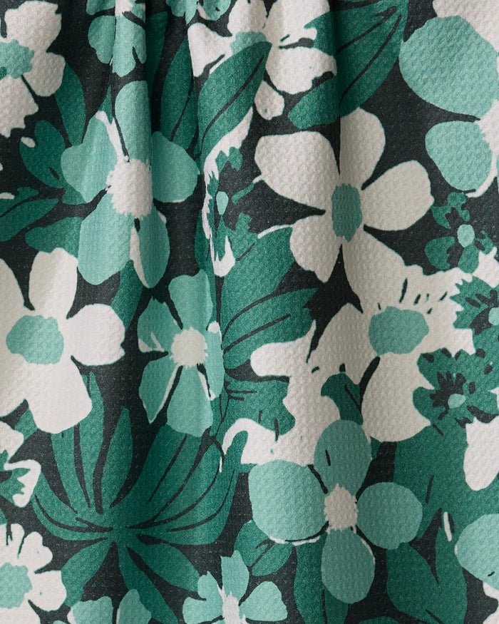 A close-up of Geometry’s Marlo Green tea towel showcases recycled fabric with white and green floral patterns and dark green leaves on a black background.