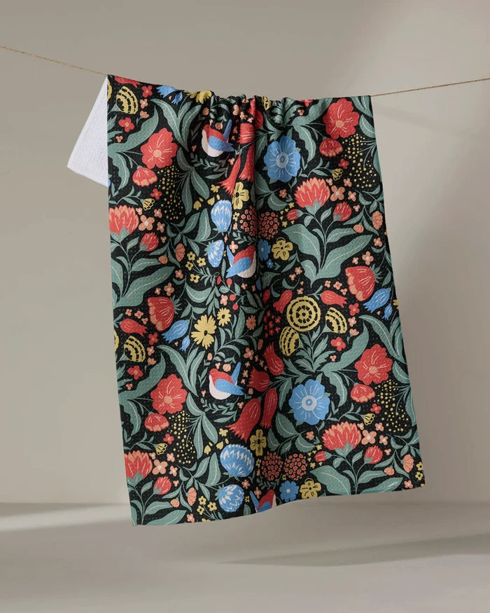 The Danelys Sidron "Marsh Wren" kitchen towel, featuring an eco-friendly design with a vibrant red, blue, yellow, and green floral pattern, hangs on a clothesline against a neutral background.