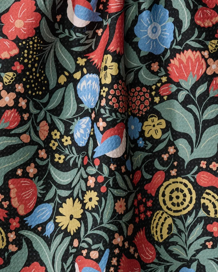 The Danelys Sidron "Marsh Wren" kitchen towel features a dense, colorful floral pattern on a black background. Eco-friendly and slightly gathered, it brings vibrant red, blue, yellow, and green flowers to brighten your kitchen space.