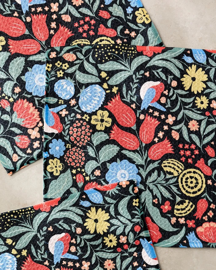 Three overlapping Marsh Wren dishtowels by Danelys Sidron, featuring a colorful floral pattern with red, blue, yellow, and green flowers on black, are arranged on a light gray surface.