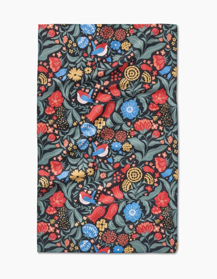 The Marsh Wren tea towel by Danelys Sidron features a vibrant floral and bird pattern with red, blue, and yellow flowers plus green leaves on a dark background, made from ultra-absorbent Geoweave fabric in a rectangular shape.