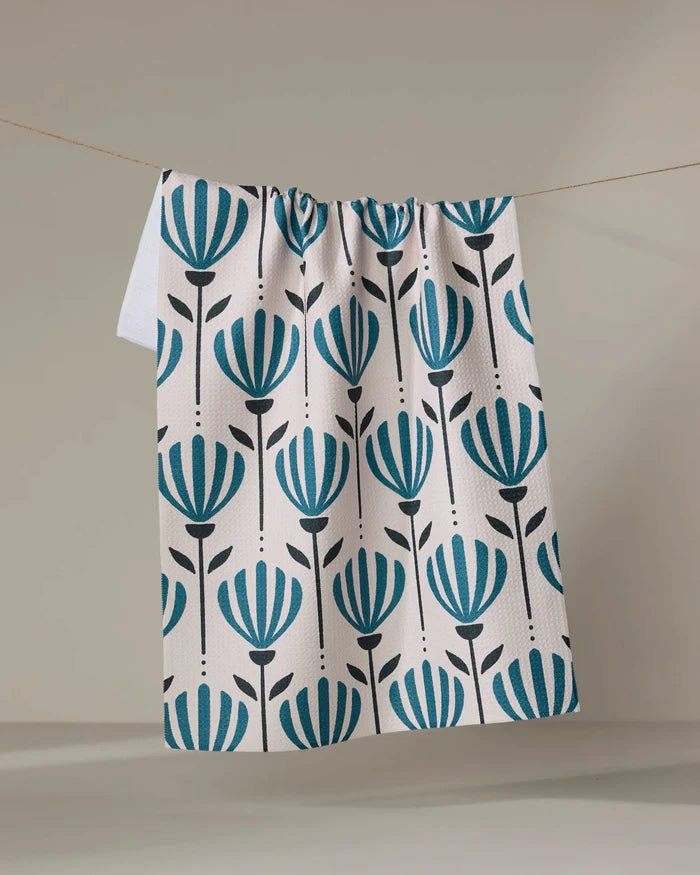 The Martine kitchen towel by Amy MacCready, made from white recycled material with a repeating blue and black stylized floral pattern, hangs on a string against a neutral background.
