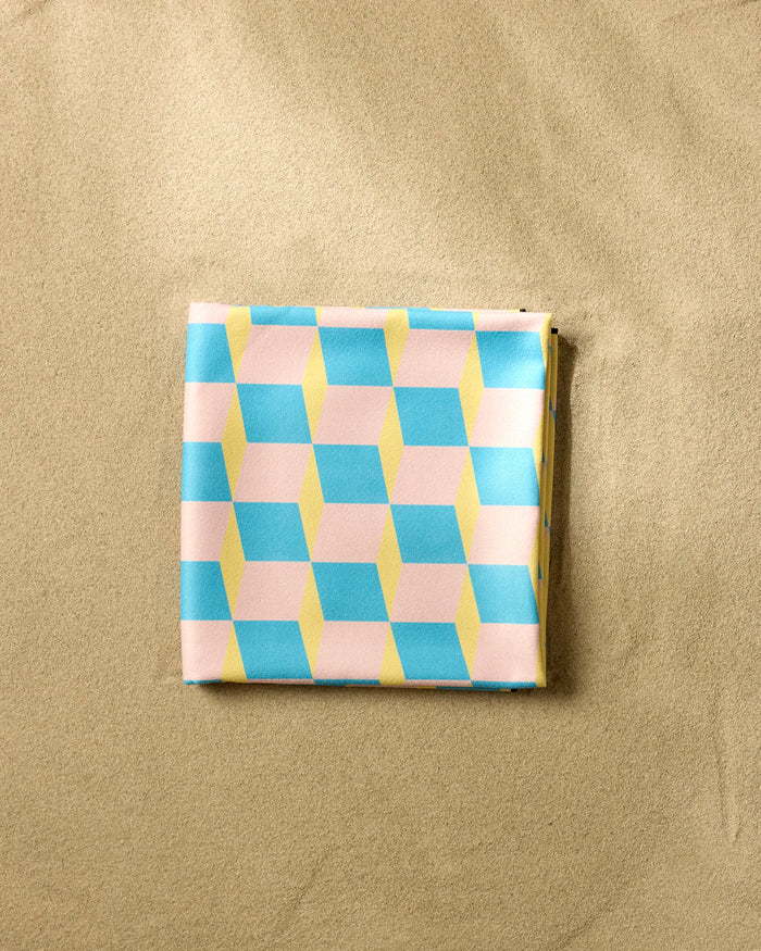 A folded GEOMETRY Math Class Micro-loop Beach Blanket featuring a pastel geometric design in blue, yellow, and pink rests on smooth, light sand.