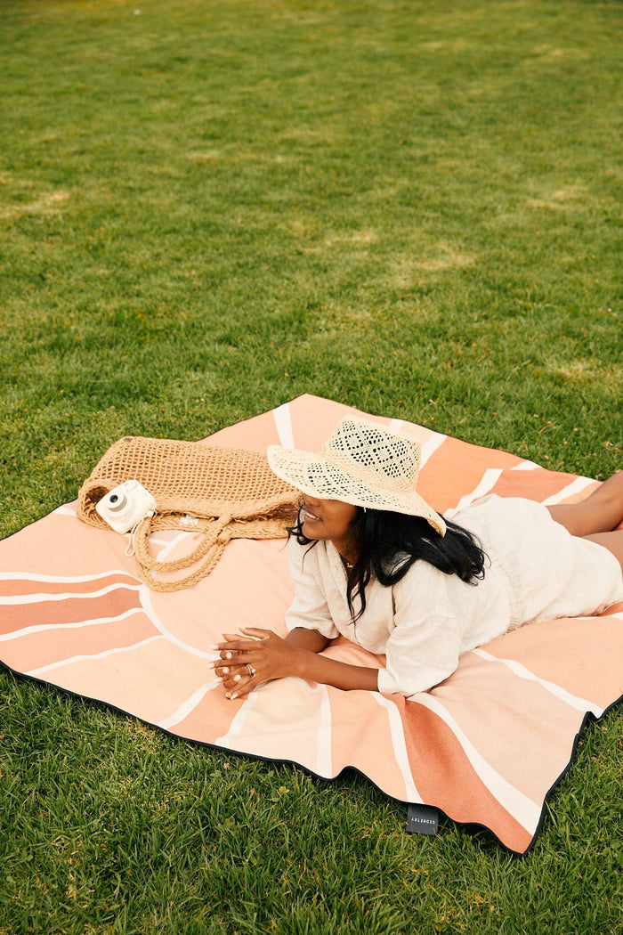 A woman in a straw hat and light outfit relaxes on her stomach, smiling, on the GEOMETRY Mauvy Moths beach blanket. Beside her are a woven bag and a small white camera as she enjoys her surroundings.