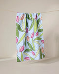 The GEOMETRY May Flowers recycled kitchen towel, featuring a pink and green tulip and leaf pattern on light blue, hangs from a string against a neutral backdrop.