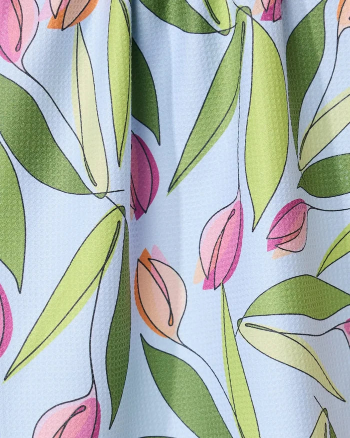 A close-up of the GEOMETRY May Flowers absorbent quick-dry towel, showcasing pink and peach tulip-style flowers with green leaves on a textured light blue background.