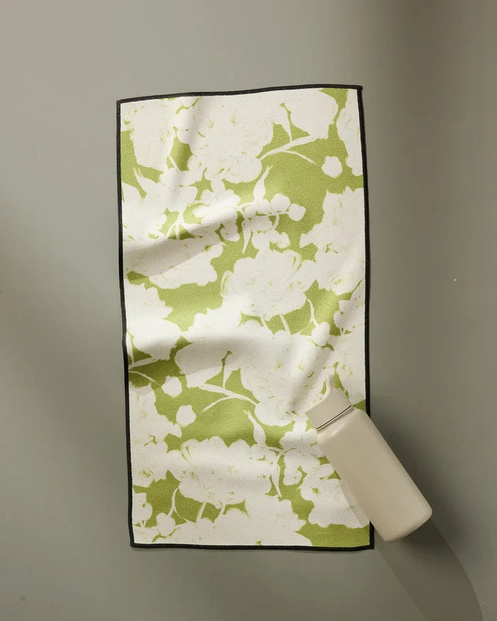 The GEOMETRY Meadow Mist Fitness Towel, featuring a green and white floral design, rests slightly wrinkled on a gray surface with a small light bottle on one corner. Its ultra-absorbent fabric combines style and practicality.