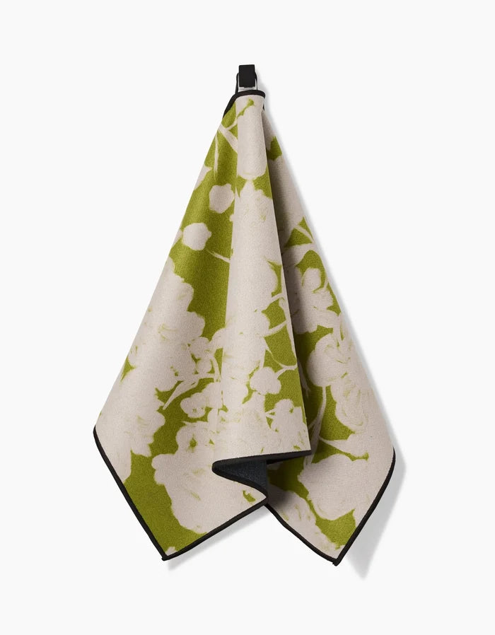 The GEOMETRY Meadow Mist fitness towel, featuring a green and cream floral design and made from recycled materials, hangs by a small black loop against a plain white background.