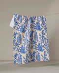 The "Mediterranean Pour" kitchen towel by Lisa Nelson, cream-colored and recycled, features blue olive oil bottles, pomegranates, and leafy branches illustrated on it, hanging against a plain beige background.