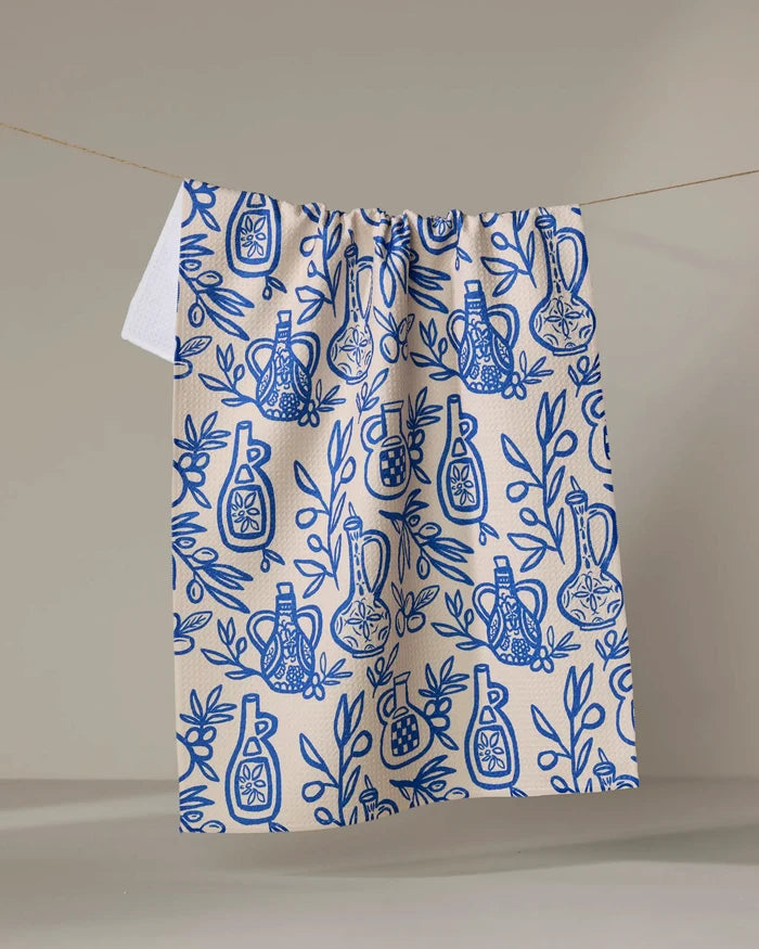 The "Mediterranean Pour" kitchen towel by Lisa Nelson, cream-colored and recycled, features blue olive oil bottles, pomegranates, and leafy branches illustrated on it, hanging against a plain beige background.