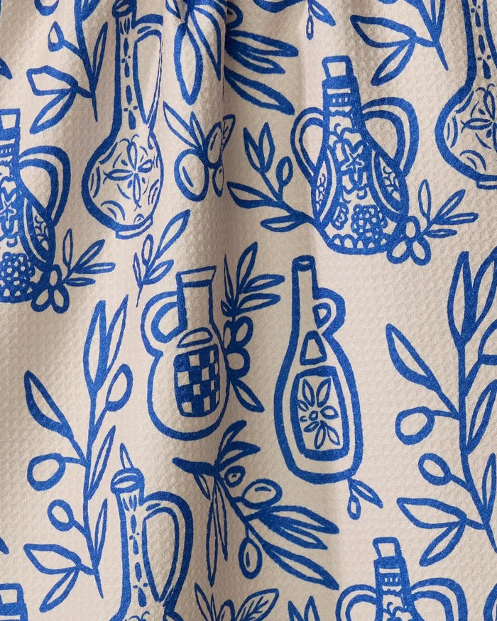A close-up of Lisa Nelson's "Mediterranean Pour" eco-friendly kitchen towel, featuring a blue and white pattern of olive oil bottles, olive branches, and leaves on a textured cream background.