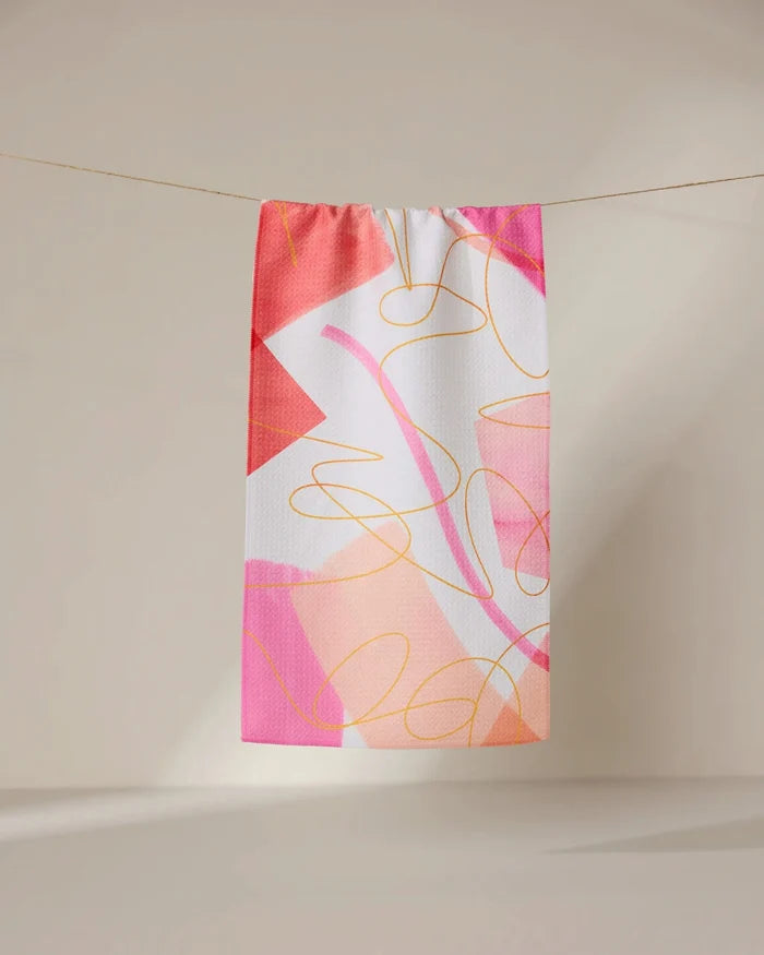 The GEOMETRY Memories towel, a rectangular ultra-absorbent piece featuring an abstract pink, orange, and white design with overlapping shapes and curved yellow lines, hangs on a clothesline against a neutral backdrop.