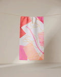 The GEOMETRY Memories towel, a rectangular ultra-absorbent piece featuring an abstract pink, orange, and white design with overlapping shapes and curved yellow lines, hangs on a clothesline against a neutral backdrop.