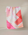 The GEOMETRY Memories recycled kitchen towel, featuring bold abstract brushstrokes and swirling lines in pink, red, orange, and yellow, hangs on a string against a neutral background.