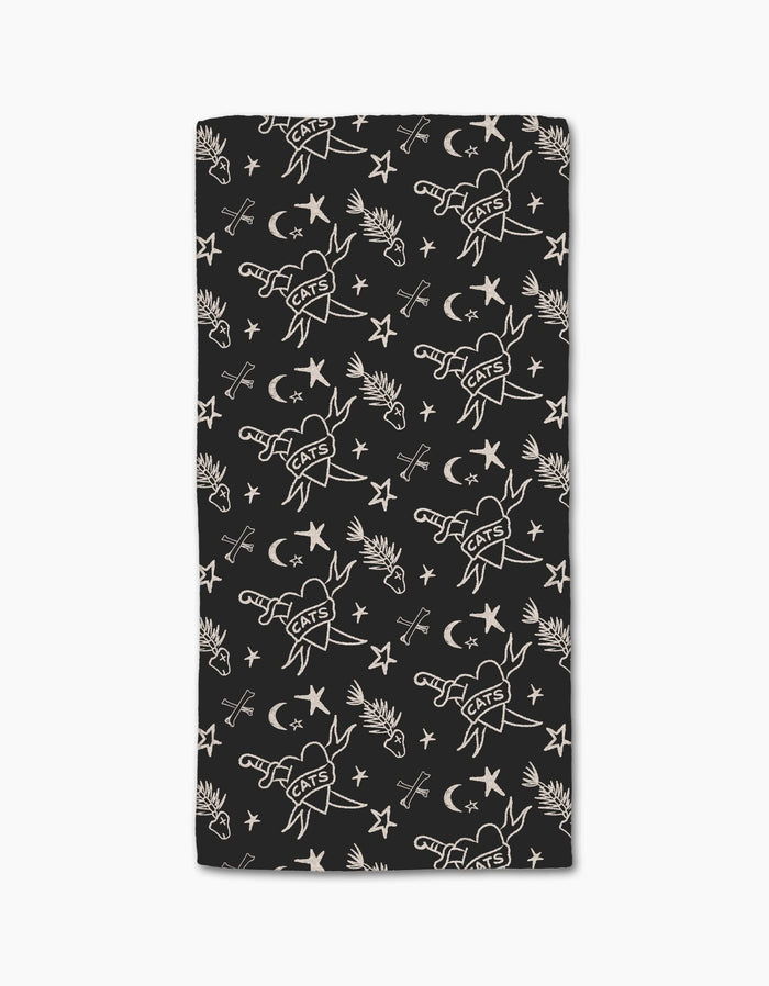 The Meow or Never bar towel by Cole Strem is a black, rectangular towel made from post-consumer recycled materials. It features white line art tattoo motifs—CATS hearts, birds, stars, moons, and flowers—on repeat.