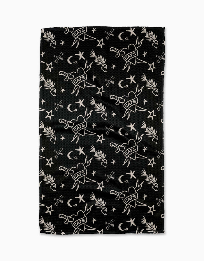 The Meow or Never Tea Towel by Cole Strem is a black Geoweave towel made from recycled materials, featuring white illustrations of swords labeled CATS, crescent moons, stars, palm leaves, and crossed arrows.