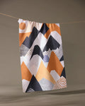 The Mesa Rise tea towel by Geometry hangs on a clothesline, showing off its vibrant abstract mountain design in orange, black, and white. Made from recycled materials, its an eco-friendly and stylish choice for your home.