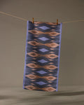 The Geometry Mesa towel, a rectangular ultra-absorbent piece with a blue, brown, and beige geometric pattern, hangs from a clothesline against a plain background, secured by two wooden clothespins.