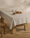A wooden table with Geometrys Metro Grid stain-resistant tablecloth in a white and black pattern holds four croissants on a plate and two white mugs. A wooden stool is beside the table on a light wooden floor.