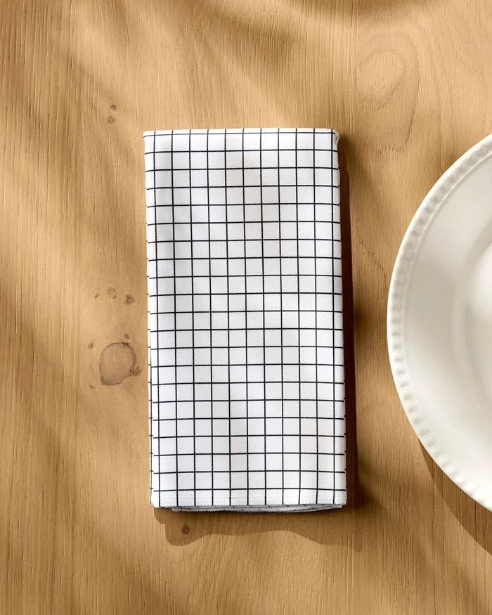 The Geometry Metro Grid reusable cloth napkin, featuring a black grid pattern, is neatly folded on a wooden table beside a white scalloped plate.