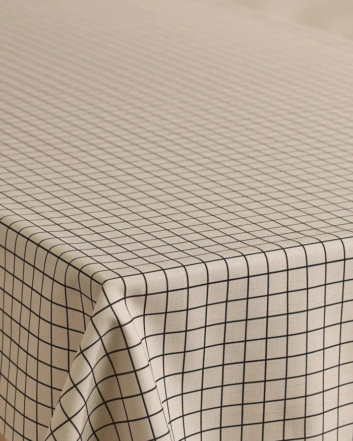 The Geometry Metro Grid tablecloth features a beige, stain-resistant fabric with evenly spaced black grid lines, draping neatly over rectangular tables for a clean, minimalistic look.