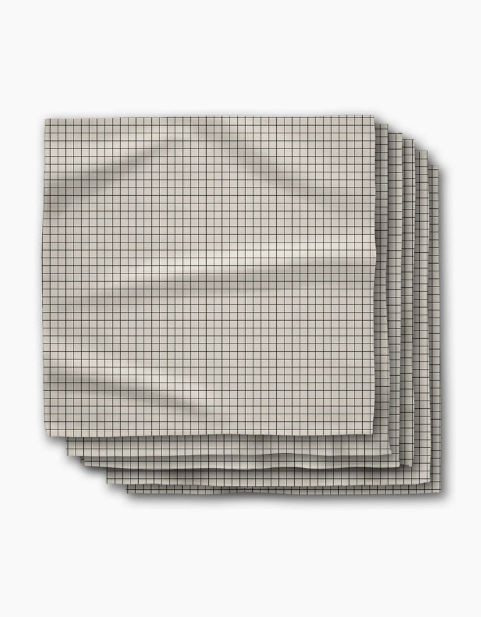 Four folded Metro Grid dinner napkins by Geometry, featuring a small black grid pattern on light beige Geosuede fabric, are stacked slightly offset on a white surface with visible creases.