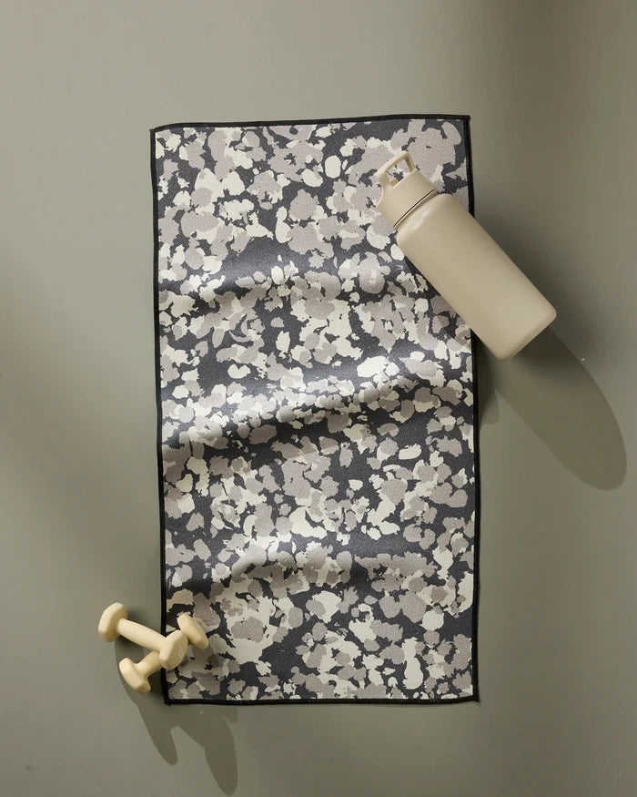 A beige water bottle and small beige dumbbells rest beside the GEOMETRY Metro Mesh ultra-absorbent black and white fitness towel on a flat, light gray surface.