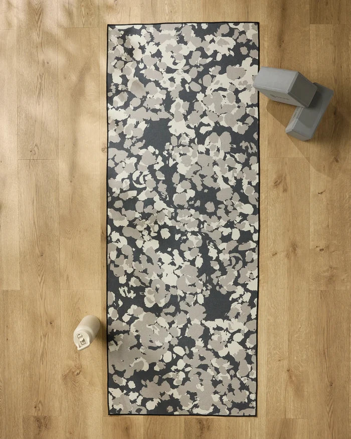The GEOMETRY Metro Mesh yoga mat with a floral pattern is laid on a wooden floor, paired with a non-slip towel, two grey yoga blocks at the top right, and a white water bottle at the bottom left.