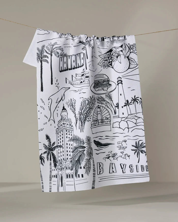 The Elisabeth Wing "Miami" kitchen towel is white, made from recycled materials, and features black line drawings of Miami landmarks, palm trees, a burger, cocktail, and the words HAVANA and BAYSIDE.
