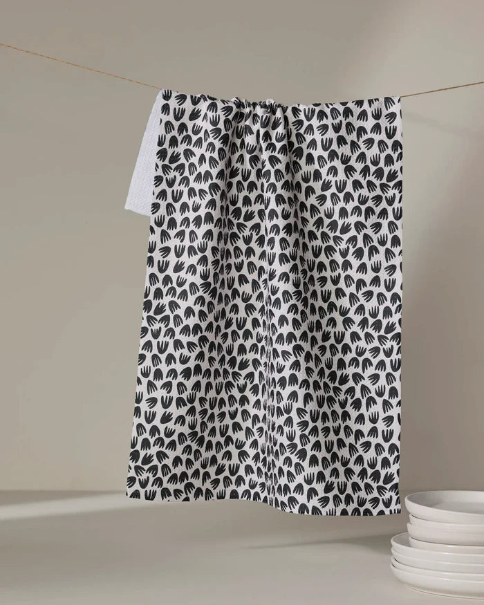 The Kayla Ann Microbloom tea towel, featuring a black handprint pattern, hangs on a clothesline beside stacks of white plates on a neutral surface.