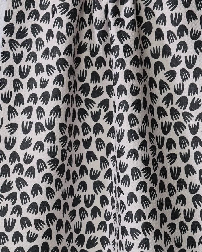 Black abstract bird-footprint shapes are scattered across the white recycled Microbloom kitchen towel by Kayla Ann, forming a repeating pattern on gently pleated fabric with subtle folds and soft shadows.