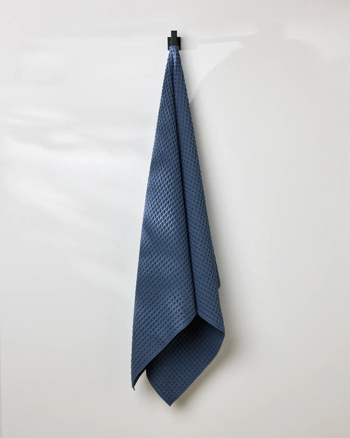 A GEOMETRY Midnight Blue bath towel hangs neatly from a black hook on a white wall, with soft light casting gentle shadows.