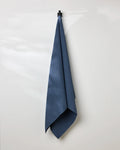 A GEOMETRY Midnight Blue bath towel hangs neatly from a black hook on a white wall, with soft light casting gentle shadows.