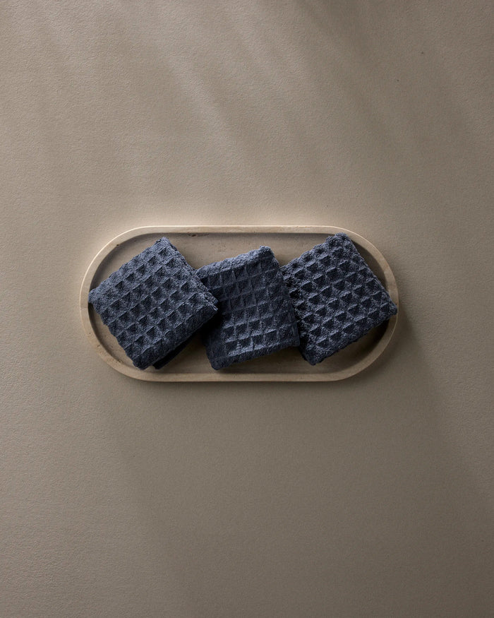 Three "Midnight Blue" waffle washcloths by GEOMETRY, made from recycled materials, are neatly displayed on a light oval tray atop a smooth taupe surface with gentle shadows.