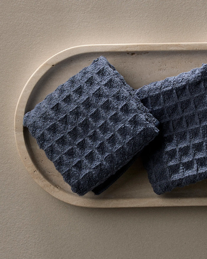 Two Midnight Blue, waffle-textured GEOMETRY towels are neatly folded and placed on an oval light stone tray against a neutral background.