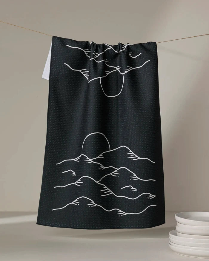 The "Midnight Mountain" tea towel by Graphics and Grain is black, made from recycled materials, and features a minimalist white line drawing of a sunrise over mountains. Absorbent and stylish, it adds a modern touch to any kitchen.