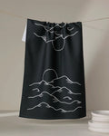 The "Midnight Mountain" tea towel by Graphics and Grain is black, made from recycled materials, and features a minimalist white line drawing of a sunrise over mountains. Absorbent and stylish, it adds a modern touch to any kitchen.