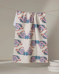 The Mimicry tea towel by Rebecca Woolbright, with vibrant butterfly wings in pink, orange, and blue, hangs on a line above a stack of white plates. The absorbent dish towel adds a pop of color to your kitchen decor.